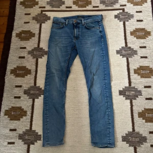 Blå skinny jeans från Nudie Jeans - Säljer ett par ljusblå skinny jeans från Nudie Jeans med klassisk femficksdesign och snygga detaljer på bakfickorna. Jeansen har orangea sömmar och är tillverkade i mjukt bomullsmaterial. Perfekt passform för dig som gillar smalare siluett.