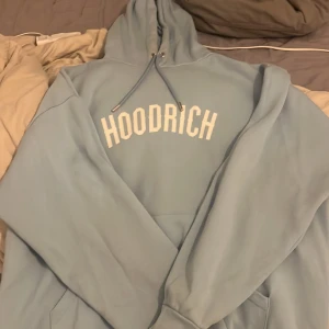 Ljusblå hoodie från Hoodrich - Säljer en ljusblå hoodie från Hoodrich i storlek L. Tröjan har vit tryckt logga på bröstet, snörning vid huvan och en klassisk känguruficka framtill. Mjuk insida och relaxed passform, perfekt för chill eller streetwear.