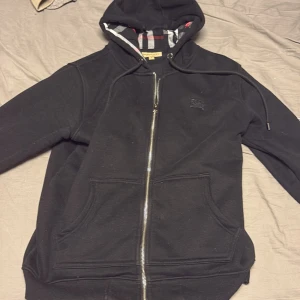 Svart Burberry hoodie  - Svart hoodie från Burberry i storlek M med klassisk rutmönstrad insida i huvan. Tröjan har hel dragkedja, snörning vid huvan och två stora fickor framtill. Tillverkad i mjuk bomull med elastan för extra komfort. Perfekt för dig som gillar stilrena designer.