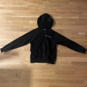 Svart hoodie från Better Bodies - Svart hoodie från Better Bodies med dragkedja och huva. Tröjan har långärmade ärmar, två fickor fram och ett litet tryck med loggan på bröstet. Perfekt för chill eller träning, mjukt material och klassisk passform.
