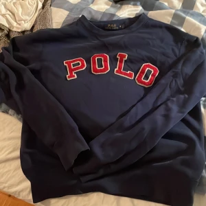 Mörkblå sweatshirt från Polo Ralph Lauren - Snygg mörkblå sweatshirt från Polo Ralph Lauren med stor röd POLO-text i broderad stil på bröstet. Tröjan har rund hals och långa ärmar, perfekt för dig som gillar klassisk streetwear med tydlig logga. Materialet är mjukt och skönt bomull.