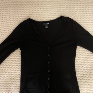 Svart kofta från H&M med fickor - Svart kofta från H&M i storlek S. Klassisk modell med v-ringning, knappar framtill och två praktiska fickor. Perfekt att ha över en t-shirt eller linne. 