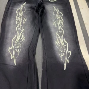 Svarta Hellstar mjukisbyxor med flames - Svarta mjukisbyxor från Hellstar med vita flames längs benen. Byxorna har elastisk midja med snörning och en bakficka. Materialet är mjukt och bekvämt, perfekt för chill dagar. Snyggt streetwear-utseende med coola detaljer framtill.