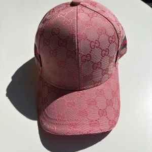 Rosa Gucci keps med GG-mönster - Snygg rosa keps från Gucci med ikoniskt GG-monogram över hela kepsen. Klassisk böjd skärm och justerbar rem bak. Sidorna har gröna och röda ränder som signaturdetalj. Tillverkad i bomull och polyester för en lyxig känsla.