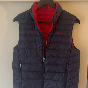 Mörkblå dunväst från Polo Ralph Lauren - Snygg mörkblå dunväst från Polo Ralph Lauren med röd insida och broderad logga på bröstet. Västen har quiltad design, två fickor med dragkedja och hög krage. Perfekt för lager-på-lager och chill stil. Dragkedja framtill för enkel på- och avtagning. Storlek: 163-174cm