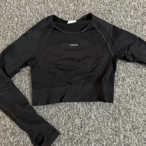 Svart croppad träningstopp Gymshark - Svart croppad långärmad träningstopp från Gymshark med ribbade muddar och tumhål i ärmarna. Toppen har öppen rygg med stor rund cut-out och Gymshark-logga baktill. Snygg, tight passform i stretchigt syntetmaterial som andas.