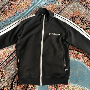 Svart Palm Angels track jacket L - Svart track jacket från Palm Angels med vita ränder längs ärmarna och dragkedja framtill. Jackan har ståkrage, två fickor med dragkedja och logga på bröstet. Tillverkad i bomull och polyester för en sportig vibe.