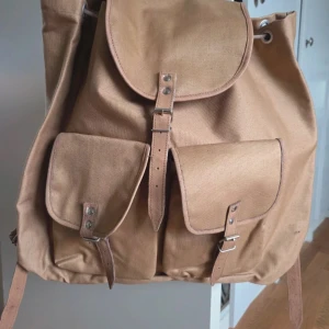 Beige canvas ryggsäck med spännen - Stilren beige ryggsäck i kraftig canvas med två stora ytterfickor framtill och snygga läderdetaljer. Väskan har justerbara axelremmar och klassiska metallspännen för en cool vintagekänsla. Perfekt för dig som vill ha en praktisk och trendig väska. Nyskick, aldrig använd. 