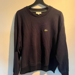 Mörkblå sweatshirt från Lacoste - Säljer en mörkblå sweatshirt från Lacoste med klassisk rund hals och det ikoniska krokodilmärket broderat på bröstet. Tröjan har en relaxed passform och är tillverkad i mjuk bomull, perfekt för chill dagar.