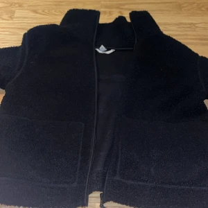 Svart teddyjacka med dragkedja - Mysig svart teddyjacka med hög krage och två stora fickor framtill. Jackan har en hel dragkedja och är tillverkad i fluffigt teddy-material som ger en cool och avslappnad vibe. Perfekt för att hålla värmen under kyliga dagar.