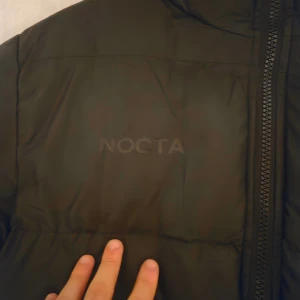 Svart NOCTA pufferjacka från Nike - Säljer en stilren och exklusiv Nike NOCTA-jacka i mycket fint skick.  Jackan är använd endast ett fåtal gånger och ser nästan ny ut. Passar både dig som har M eller L, då passformen är flexibel och bekväm.  🟩 Perfekt för både vardag och streetwear-stil!  🟦 Pris kan diskuteras vid snabb och smidig affär.
