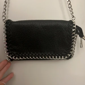 Svart axelväska med kedjedetaljer - Cool svart axelväska i skinnimitation med grov silverfärgad kedja runt kanterna och som axelrem. Väskan har lock med magnetlås och dragkedja på insidan. Inte rostad 