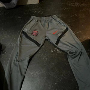 Grå PSG träningsbyxor från Nike S - Säljer ett par grå träningsbyxor från Nike med PSG-logga och röd swoosh på framsidan. Byxorna har elastisk midja, svarta detaljer längs benen och är tillverkade i mjukt syntetmaterial. Perfekta för dig som gillar sportig stil och fotboll.