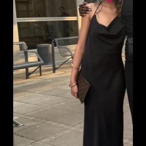 Säljer min balklänning  - Svart långklänning med tunna axelband och snygg draperad urringning. Klänningen har en öppen rygg och faller rakt ner i en elegant siluett. Materialet är slätt och ger en lyxig känsla, perfekt för dig som vill sticka ut.