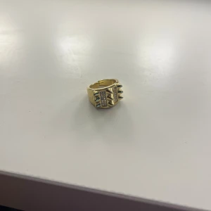 Guldfärgad ring med nitar och stenar - Cool bred guldfärgad ring med två rader av små glittriga stenar och markerade nitar på framsidan. Ringen har en chunky design som sticker ut och ger en edgy vibe. Perfekt för dig som gillar att addera lite attityd till din stil.