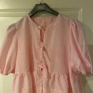 Rutig rosa kortklänning med knyt - Supersöt rosa kortklänning med små vita rutor och puffiga ärmar. Klänningen har tre knyt framtill och en lös, luftig passform. Perfekt för dig som gillar gulliga detaljer och en romantisk vibe.