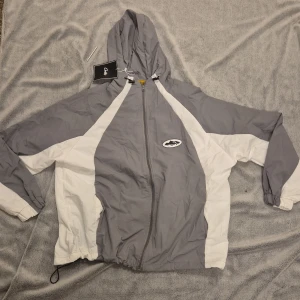 Grå och vit vindjacka från corteiz storlek s - Snygg vindjacka från corteizi grått och vitt med huva och dragkedja. Jackan har coolt tryck på bröstet och meshfoder på insidan. Elastiska muddar och snörning nedtill för extra passform. Perfekt för dig som gillar streetwear och sportig stil.