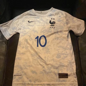 Frankrike Mbappé fotbollströja Nike - Säljer en officiell Frankrike landslagströja från Nike med Mbappé och nummer 10 på ryggen. Tröjan är vit med ljusblå mönster och har korta ärmar. Tillverkad i lätt och ventilerande material (VaporKnit) med FFF-logga och två stjärnor på bröstet.