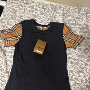 Burberry t-shirt med rutiga ärmar - Svart Burberry t-shirt i bomull med klassiska rutiga kortärmar i beige, rött och svart. Snygg kontrast mellan den enfärgade kroppen och de ikoniska ärmarna. Perfekt för dig som gillar designerplagg med unik touch. Helt ny och strlk L i barnstrlk