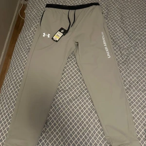 Grå träningsbyxor Under Armour  - Snygga grå träningsbyxor från Under Armour med svart dragsko i midjan och logga på benet. Byxorna har en loose passform och är gjorda i lätt syntetmaterial som andas. Perfekta för gymmet eller chill hemma. Strlk L men passar även M. Kan gå ner nån slant vid snabb affär vill få bort dom.