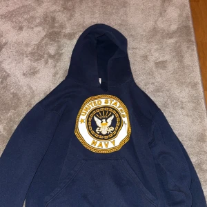 Mörkblå United States Navy hoodie - Säljer en mörkblå hoodie med stort United States Navy-emblem på bröstet och tydlig NAVY-text på ryggen. Tröjan har huva och magficka, samt detaljer i vitt och gult. Perfekt för dig som gillar militärstil och vill sticka ut.