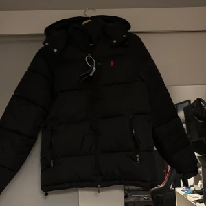Svart pufferjacka från Polo Ralph Lauren - Svart pufferjacka från Polo Ralph Lauren med huva, två stora fickor med dragkedja och röd broderad logga på bröstet. Jackan har quiltad design, justerbar nederkant och är helfodrad med blank insida. Perfekt för kalla dagar och har en clean, modern look.