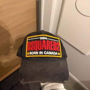 Svart DSQUARED2 keps med brodyr - Svart keps från DSQUARED2 med stor broderad logga och texten 'Born in Canada' framtill i rött, vitt och gult. Baksidan har röd brodyr med 'Born to be a fighter' och '1995'. Justerbar passform och tillverkad i bomull. Perfekt för dig som gillar streetwear och statement-plagg.