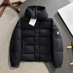 Svart Moncler pufferjacka med huva - Svart pufferjacka från Moncler med stor huva och snörning, ikonisk logga på ärmen och praktiska fickor. Jackan har både dragkedja och knappar framtill, samt quiltad design som ger en schysst look. Perfekt för kalla dagar när du vill vara både varm och stilren.