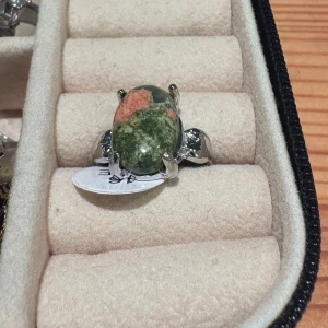 Silverring med grön och rosa sten - Unik silverring i storlek 16 med en oval, polerad sten i gröna och rosa nyanser. Ringen har en bred, blank silverfattning som håller stenen stadigt på plats. Perfekt för dig som gillar statement-smycken med naturliga färger och cool design.