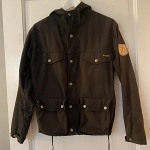 Svart vindjacka från Fjällräven G-1000 - Svart vindjacka från Fjällräven i G-1000-material med huva, fyra fickor med tryckknappar och dragsko i midjan. Klassisk Fjällräven-logga på ärmen och snygga detaljer i metall. Perfekt för dig som gillar outdoor och streetstyle.