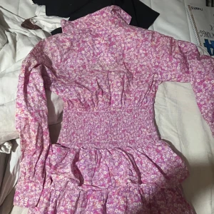 Ljusrosa blommig klänning Pernilla Wahlgren - Superfin ljusrosa kortklänning från Pernilla Wahlgren med blommigt mönster, volanger och smock i midjan. Klänningen har långärmade puffärmar, knappar framtill och en klassisk krage. Perfekt för dig som gillar romantisk och feminin stil.