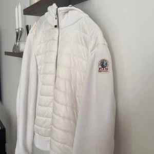 Vit quiltad pufferjacka från Parajumpers - Snygg vit pufferjacka från Parajumpers med huva och quiltad framsida och rygg. Jackan har dragkedja framtill och ett broderat märke på ärmen. Materialmix med polyester och bomull, perfekt för dig som gillar stilren och sportig look. Storleks lappen är avklippt. Men QR-koden lappen är kvar som tar dig till Parajumpers hemsida genom att scanna den. 