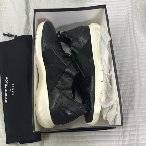 Svarta sneakers Hypnotic Yellow x Lynk & Co - Snygga svarta sneakers från Hypnotic Yellow för Lynk & Co med chunky vit sula och detaljer i skinn och textil. Skorna har elastiska band över foten och en modern, sportig look. Perfekta för dig som gillar stilrena och bekväma sneakers med unik design.