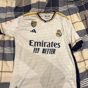 Real Madrid Vini Jr. 7 matchtröja - Säljer en officiell Real Madrid fotbollströja från Adidas med Vini Jr. och nummer 7 på ryggen. Tröjan är vit med tunna ränder, guld- och marinblå detaljer, samt klubbmärke och FIFA-märke på bröstet. Materialet är lätt och andas, perfekt för träning eller match.