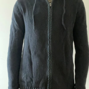 Svart stickad hoodie Adrian Hammond - Svart stickad hoodie från Adrian Hammond med dragkedja framtill och snörning i huvan. Långärmad modell med broderad AH-logga på nedre delen. Perfekt för dig som gillar stilren och enkel design med lite edge.