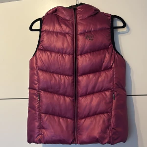 Mörkrosa dunväst från Kingsland - Snygg mörkrosa dunväst från Kingsland med glansig finish och broderad logga på bröstet och ryggen. Västen har dragkedja framtill, två fickor med dragkedja och en hög krage med huva. Perfekt för dig som gillar ridning och vill ha en sportig look. Västen har inte alls varit använd den har ingen prislapp för jag trodde jag skulle använda den men nu är den bara kvar i garderoben.