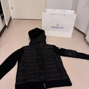 Svart Moncler pufferjacka med huva - Svart pufferjacka från Moncler med huva och ribbstickade ärmar. Jackan har dragkedja framtill, två fickor med dragkedja och en extra ficka med dragkedja på ärmen. Moncler-logga på vänster ärm. Snygg, stilren och perfekt för kyliga dagar.