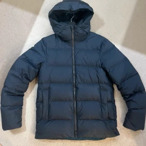 Mörkblå dunjacka från J.Lindeberg - Snygg mörkblå dunjacka från J.Lindeberg med huva och stilren design. Jackan har en quiltad look, dragkedja framtill och diskret logga på bröstet. Perfekt för kalla dagar och enkel att matcha med jeans eller hoodie.