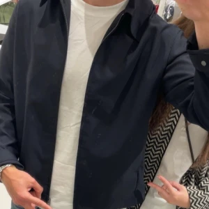 Mörkblå overshirt med dragkedja - Snygg mörkblå overshirt med klassisk krage och rak passform. Jackan har dragkedja framtill och långa ärmar, perfekt att slänga på över en t-shirt eller hoodie. Materialet är slätt och känns lätt, vilket gör den enkel att styla med jeans eller chinos.