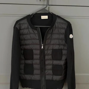 Svart pufferjacka från Moncler - Svart pufferjacka från Moncler med stickade ärmar och quiltad front. Jackan har två stora fickor framtill, rund hals och dragkedja hela vägen. Moncler-logga på vänster ärm. Perfekt för dig som vill ha en snygg och stilren jacka med exklusiv känsla.