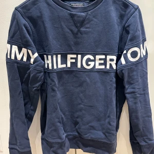 Marinblå sweatshirt Tommy Hilfiger - Snygg marinblå sweatshirt från Tommy Hilfiger med stort vitt logotryck runt hela bröstet och armarna. Tröjan har rund hals, långa ärmar och ribbade muddar vid ärmslut och nederkant. Perfekt för dig som gillar klassisk streetstyle.