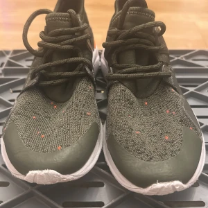 Gröna sneakers med orange detaljer - Snygga sneakers i mörkgrönt med stickad ovandel och små orange prickar. Skorna har vita, mönstrade sulor och coola orange detaljer vid hälen. Ovandelen är i textil och syntet, med snörning framtill för en sportig look. Perfekta för dig som gillar streetstyle.