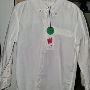Vit skjorta från Lindex, stl 122 - Klassisk vit skjorta från Lindex i storlek 122. Skjortan har lång ärm, knappar framtill och en bröstficka. Perfekt för en clean och stilren look. Tillverkad i mjuk bomull för skön känsla hela dagen.