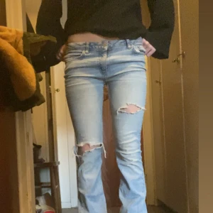 Ljusblå ripped jeans med bootcut - Säljer ett par ljusblå jeans med snygga slitningar på båda knäna och bootcut-snitt. Jeansen har klassisk femficksdesign och låg midja, perfekta för en chill och trendig look. Lite korsta i benen 