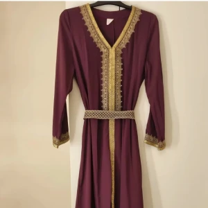 Lång vinröd klänning med gulddetaljer - Elegant långklänning i vinröd färg med vackra guldfärgade spetsdetaljer längs v-ringningen, ärmsluten och nederkanten. Klänningen har lång ärm och medföljande bälte i matchande tyg och mönster. Perfekt för dig som vill sticka ut med en klassisk och lyxig look.