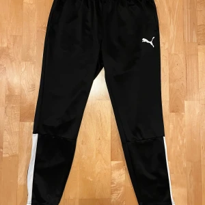 Puma Träningsbyxor Teamliga - Svart/Vit, strl L - Svarta träningsbyxor från Puma med vit logga på höften och vita paneler längs benen. Dragkedja finns vid benöppningen och vid byxfickorna. Drycell. Storlek L.