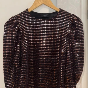 Glittrig svart/rosa topp “Sisters Point” - Supersnygg svart topp från Sisters Point i storlek XS. Toppen har glittriga paljetter i rader över hela tyget, puffiga axlar och korta ärm. Materialet är en mix av nylon, metallisk tråd och elastan som ger en stretchig och bekväm passform. Perfekt för att sticka ut på festen!