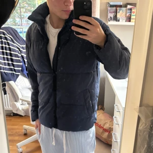 Mörkblå pufferjacka med dragkedja - Säljer en mörkblå pufferjacka med hög krage och dragkedja framtill. Jackan har en clean look, två sidofickor och är perfekt för kyligare dagar. Passformen är normal och jackan är vadderad för extra värme. Märke ej synligt.