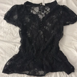 Svart spetsblus med knappar - Säljer en svart blus i  spets med blommigt mönster. Blusen har korta ärmar, v-ringning och dekorativa knappar framtill. Den sitter figurnära, har köpt secondhand så de står inte vart den är köpt💕 i storlek 36/ S