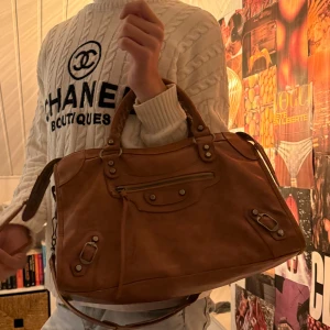 Brun Balenciaga handväska i skinn - Säljer en brun handväska från Balenciaga i äkta skinn med coola metalldetaljer och dragkedja framtill. Väskan har både handtag och axelrem, perfekt storlek för allt du behöver. Snygg design med klassiska Balenciaga-nitar och platta med logga inuti. Lägg gärna bud!